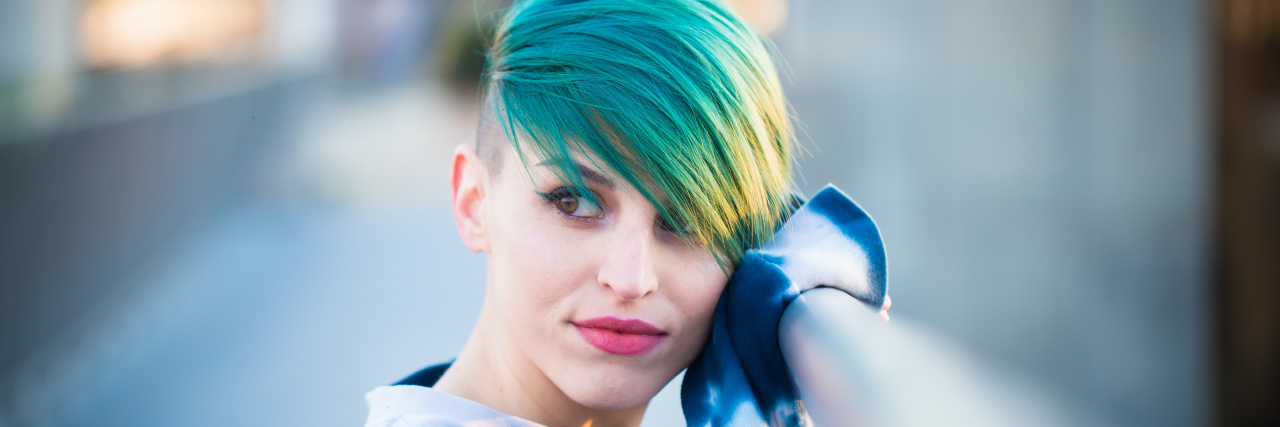The Gaps in Mental Health Care Hospitals Young woman with short green hair in a tie-dyed shirt standing outside looking away