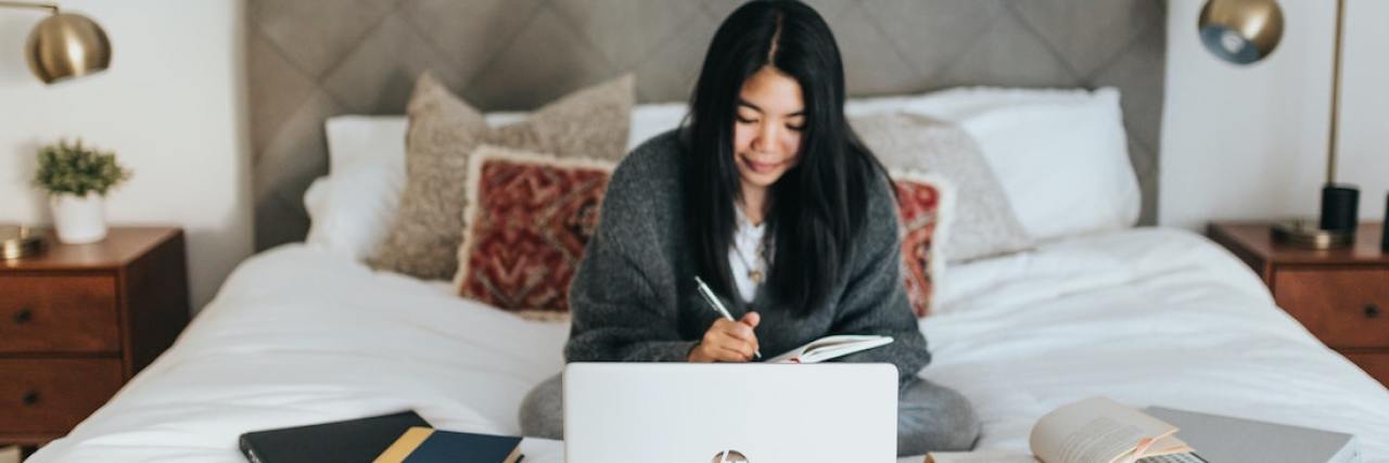 A woman working in bed, looking at a laptop. There are books surrounding her.