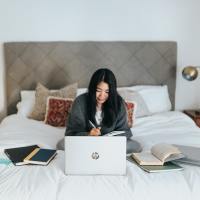 A woman working in bed, looking at a laptop. There are books surrounding her.