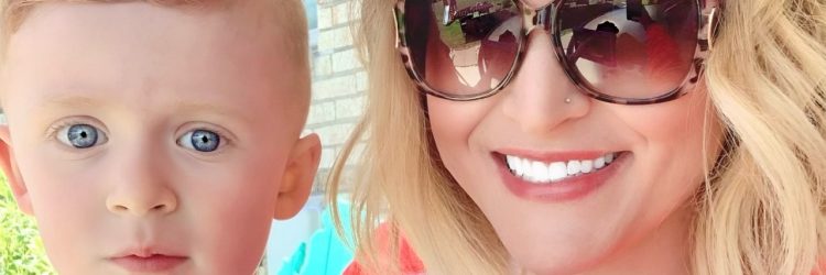 A mother and their austic child smiling and looking at the camera. The mother has blonde hair and brown sunglasses with a pink brown lipstick. she's also wearing a coral colored shirt. 