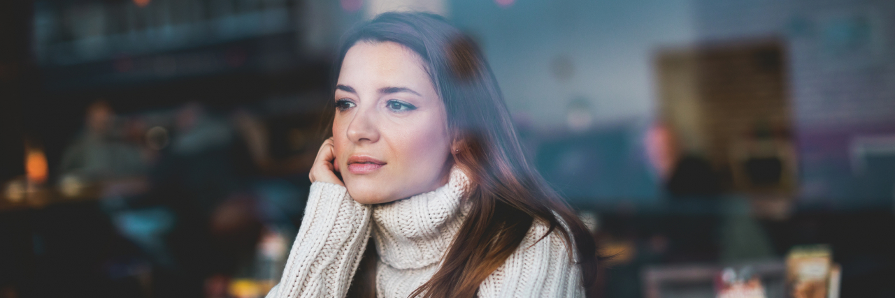 How the Meaning of Mental Illness Has Changed With Increased Exposure Young woman in a white sweater sitting by herself in a cafe