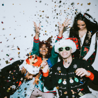 A diverse group of people wearing Christmas and holiday sweaters, santa hats and sunglasses throw confetti in the air