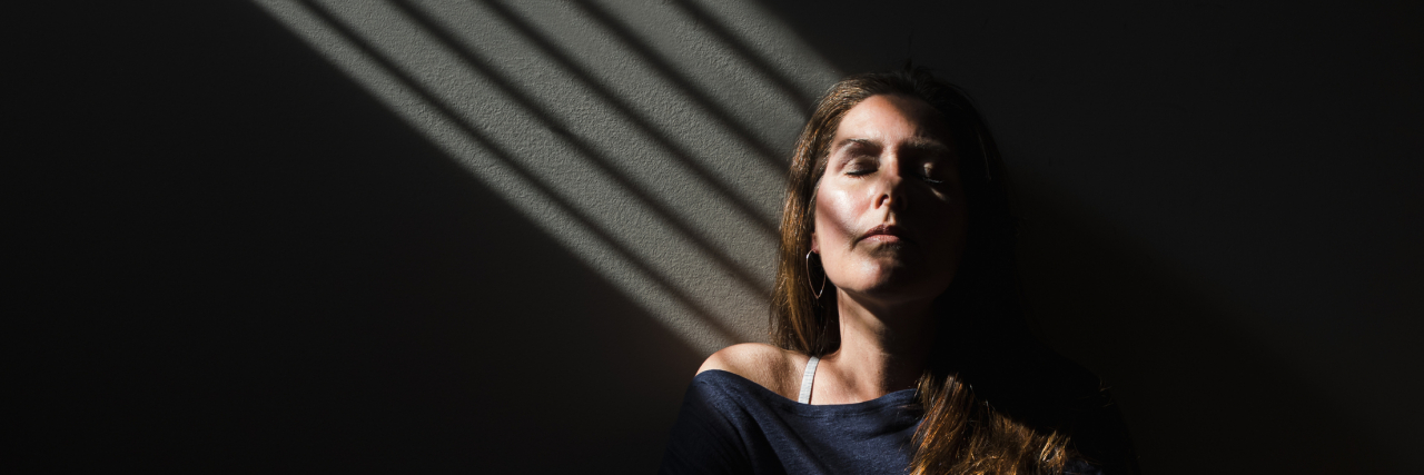 5 Ways I Stay Positive Living With Epilepsy Portrait of a woman sitting in a patch of light with her eyes closed, surrounded by darkness