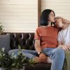 Tattooed lesbians in brown and white t-shirts kiss and hug sitting on leather sofa at home with white wall