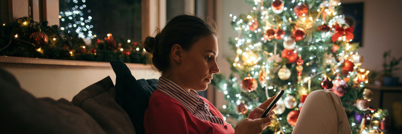 Woman sitting alone on sofa next to a decorated Christmas tree and using phone