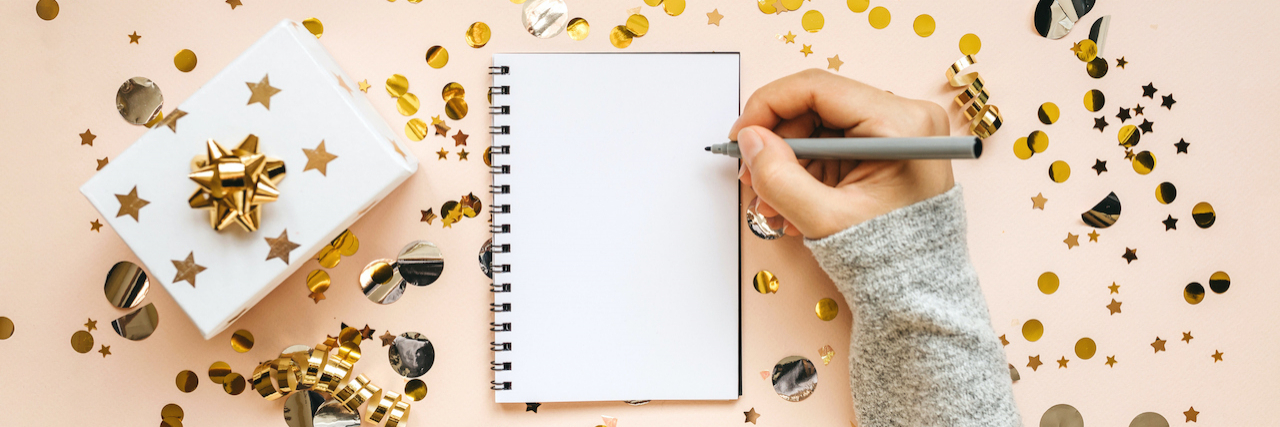 Hand holding pen above blank notebook, surrounded by gold confetti