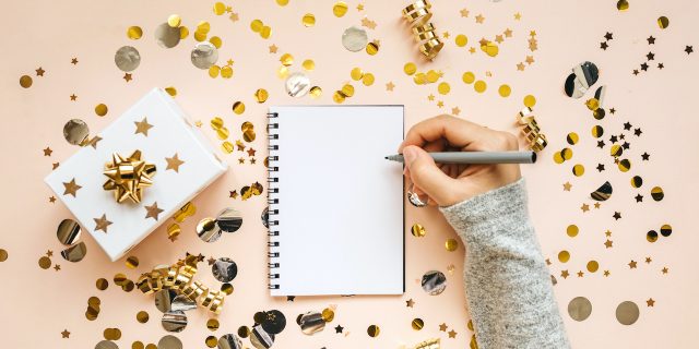 Hand holding pen above blank notebook, surrounded by gold confetti