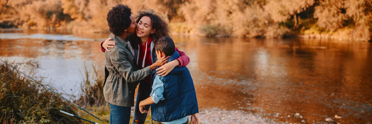 Black mother hugging her smiling children by river on fall day