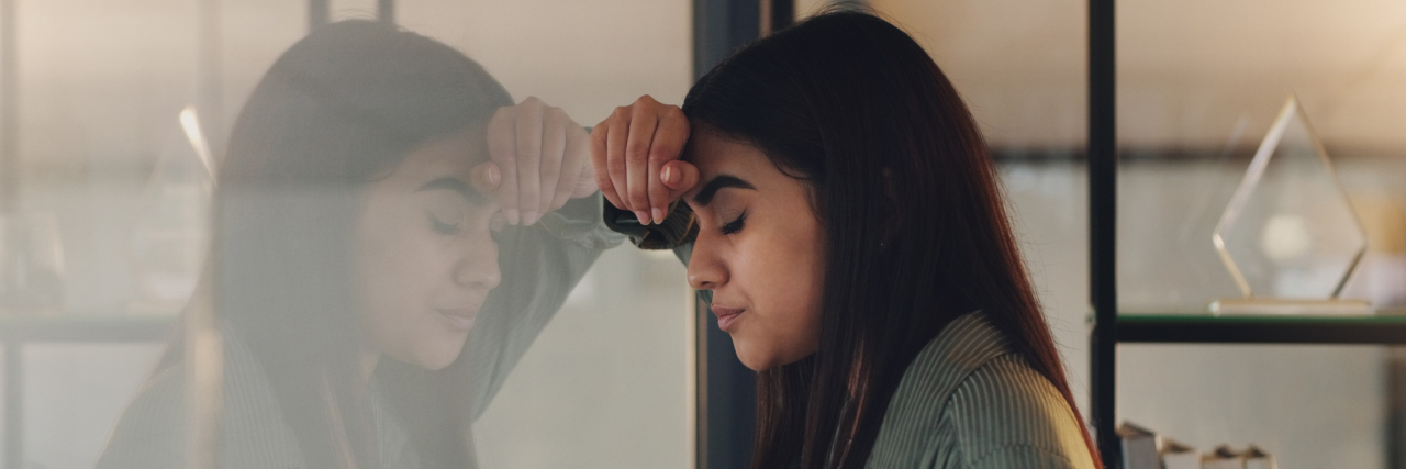 How Burnout Impacts My Ability to Manage Constant Suicidal Thoughts A young woman with her head against the wall and eyes closed, stressed