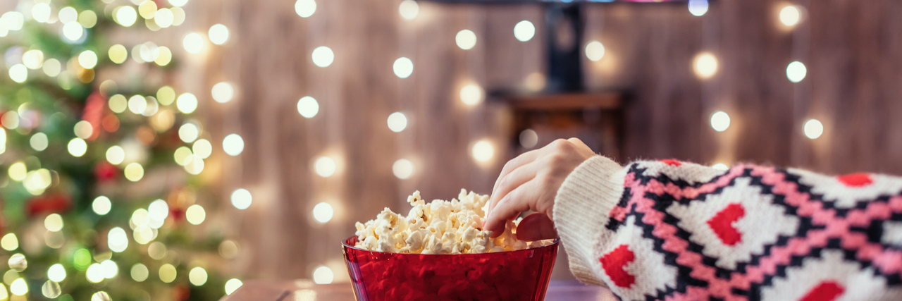 Tips For Weathering the Holiday Blues and Enjoying This Season Hand reaching for popcorn bowl in front of TV that is next to Christmas tree and holiday lights