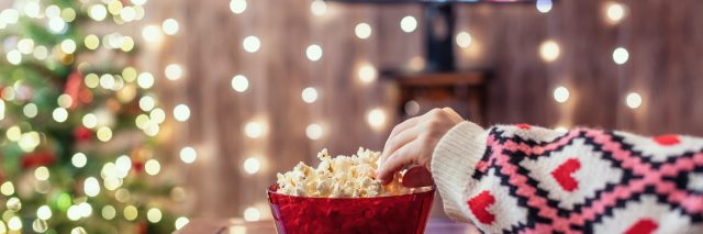 Hand reaching for popcorn bowl in front of TV that is next to Christmas tree and holiday lights