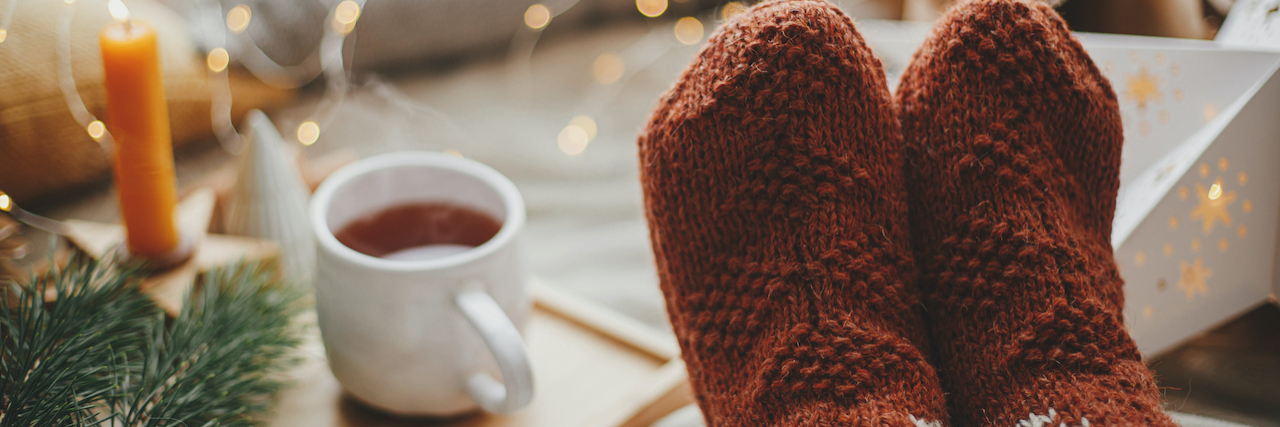 Making the Most of Spending Holidays At Home With Rare Disease Woolen socks on bed with cup of tea, golden lights, trees, candle and pillows.