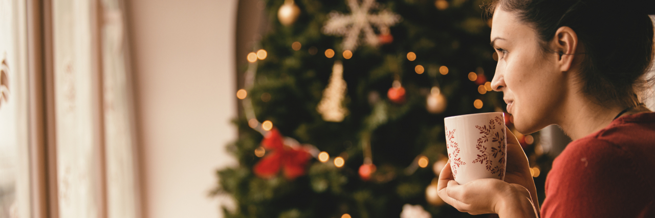 4 Ways to Manage Holiday Stress and Your Mental Health Young woman drinking tea by the Christmas tree, looking through window.