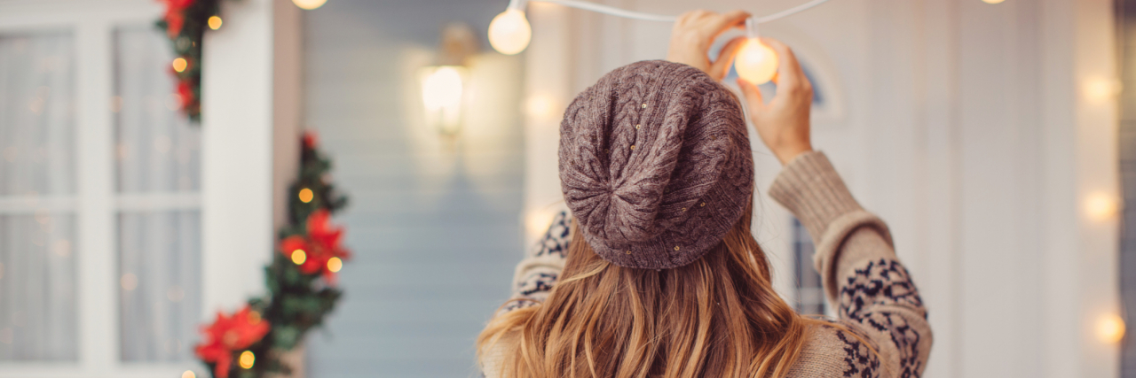 Coping With Holiday Mental Health Triggers During the Holiday Season Back of a young woman in a sweater putting up string lights in a room decorated for Christmas