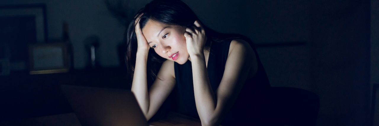 Asian woman sitting in the dark with head in her hands, her face illuminated by the light from a laptop