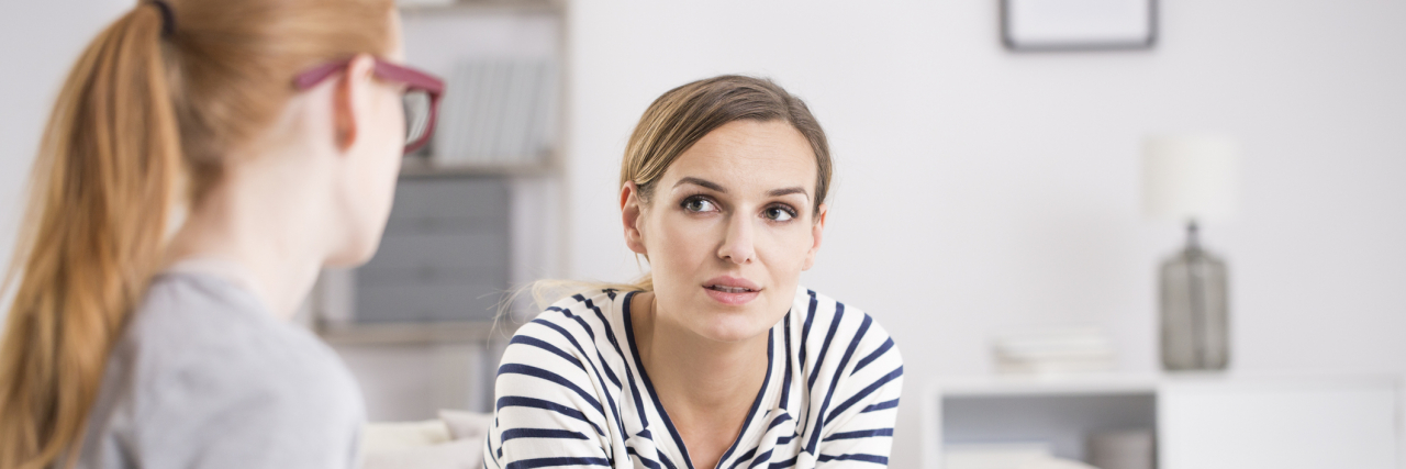 Realizing My Therapist Is Human Too Helped Challenge My Perfectionism A woman in glasses with a clipboard listening to a woman across from her on the couch talk; therapy