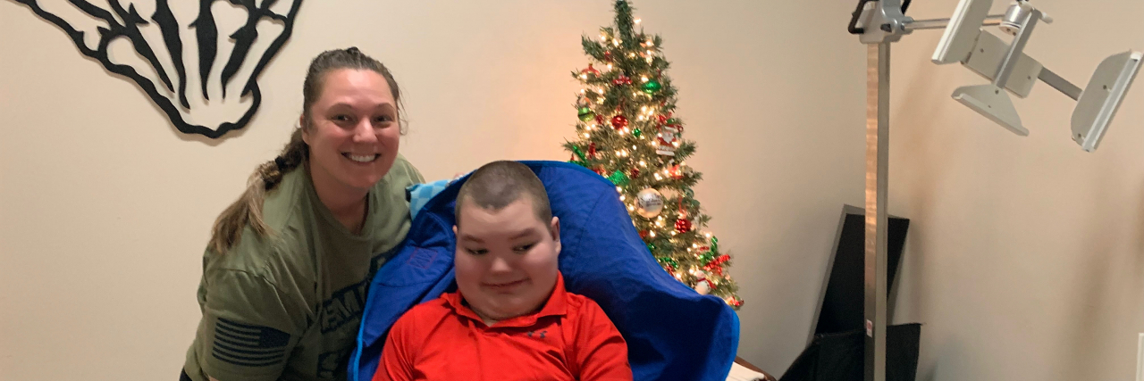 When Our Family Found the Holiday Spirit During a Hospital Visit Jacob sitting in his chair with his Mom posing next to him in front of a Christmas tree