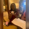 The edges of the pictures are blurred out. Centered you have a Black girl in a fabulous maroon jumpsuit with bows on the sleeves blowing out a birthday cake in a ritzy hotel in Washington DC. Her hair is straightened and next to her on the couch there's a pink mask and a camera. A pink dinner napkin sits perfectly on her lap.