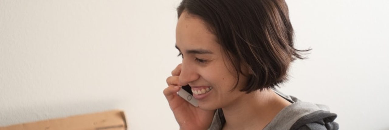 How Dialectical Behavior Therapy 'Skills Coaching' Helps Me Learn to Accept Help A person with short brown hair and brown eyes wearing a gray hoodie smiles while sitting on a bed and speaking on a cell phone.