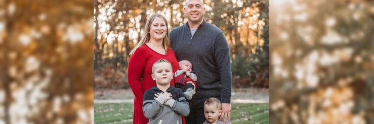 A family of five. A mother in a red dress stands to the left, mirrored by her husband to the right in a gray sweatshirt with a lighter gray undershirt, an infant betwen them and two sons, one with his arms crossed over his chest and the other looking off camera.