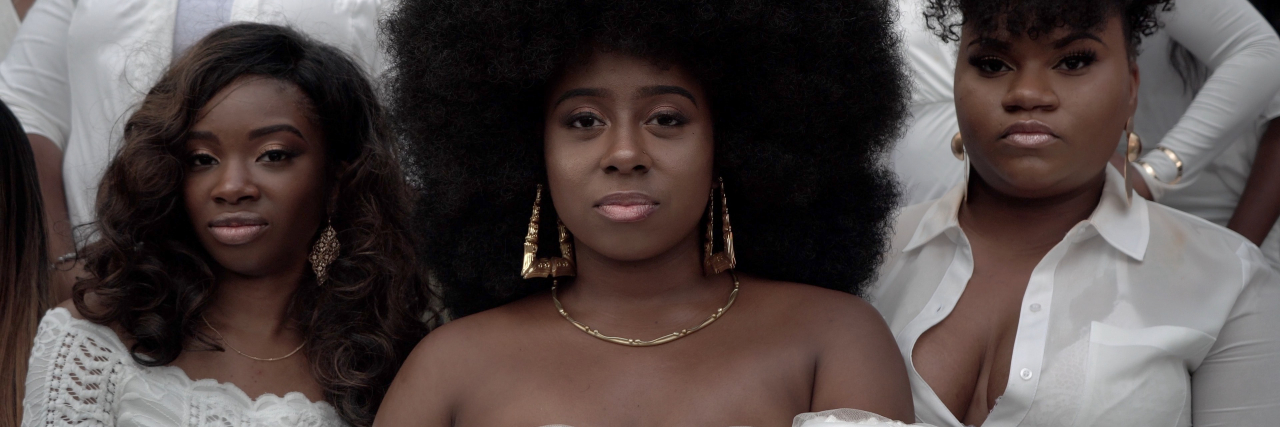 A group of beautiful Black woman staring into the camera, all dress in white. The one in the middle has an afro and gold earrings, and she's flanked by another woman with a pompadour up-do and one wearing her hair wavy and down her back.