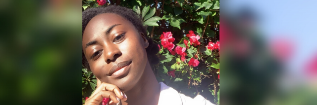 A beautiful photo of the writer. Her dark skin is practically glowing. She rests her head on her hand and is wearing a pink polo. Behind her is a big flower bush.