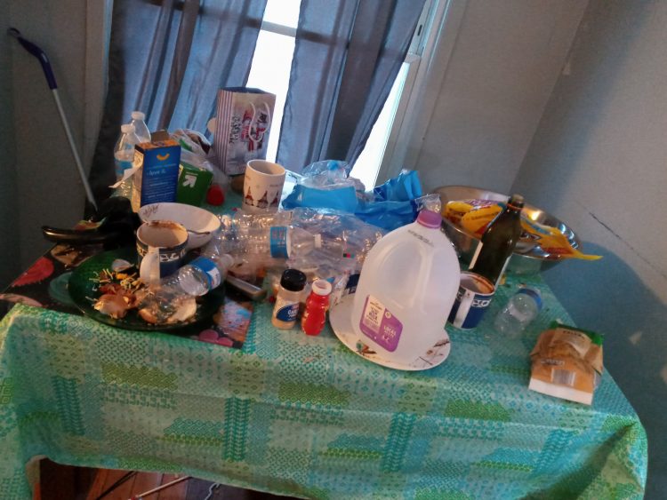 A messy kitchen table with food, dishes, old jars, and more everywhere. There's a blue table cloth underneath.