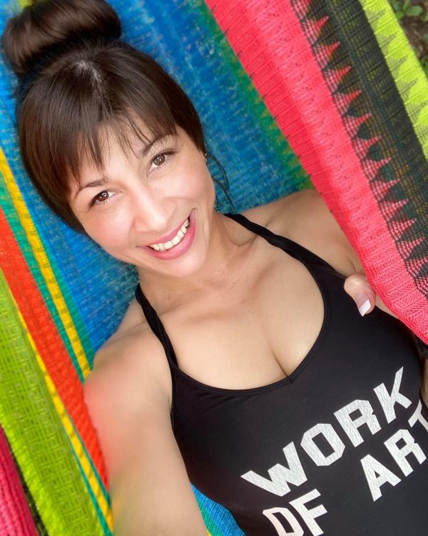 A woman with brown hair and brown eyes wearing a black tank top with the phrase "Work of Art" written on it in white smiles.
