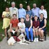 Woman in a yellow dress with bouquet of pink roses with man and four adult children, five grandchildren, and one golden lab service dog pose for post-wedding photo in a park
