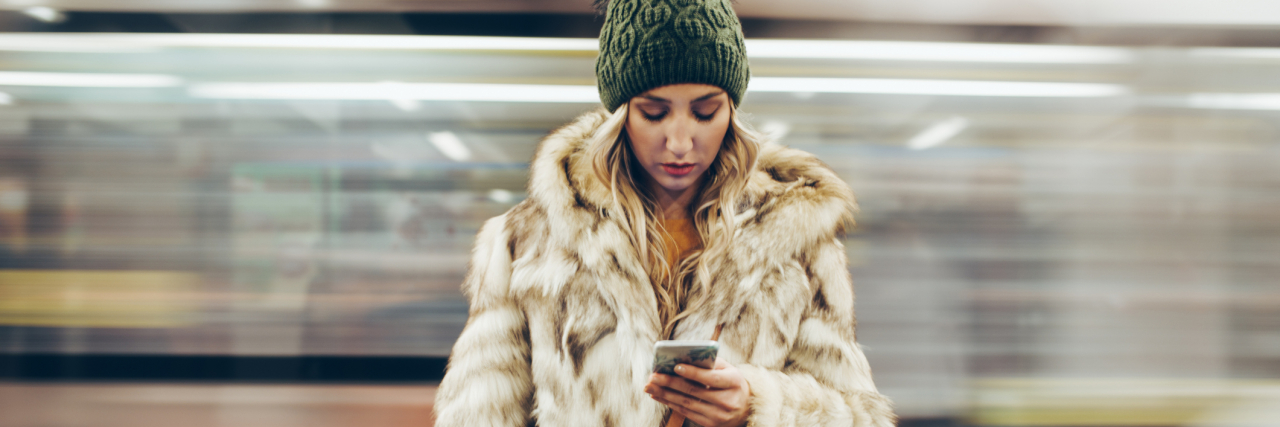 How to Avoid Anxiety on Social Media (and Not Cause It) Young woman in a fur coat looking at her phone with a blurred train behind her