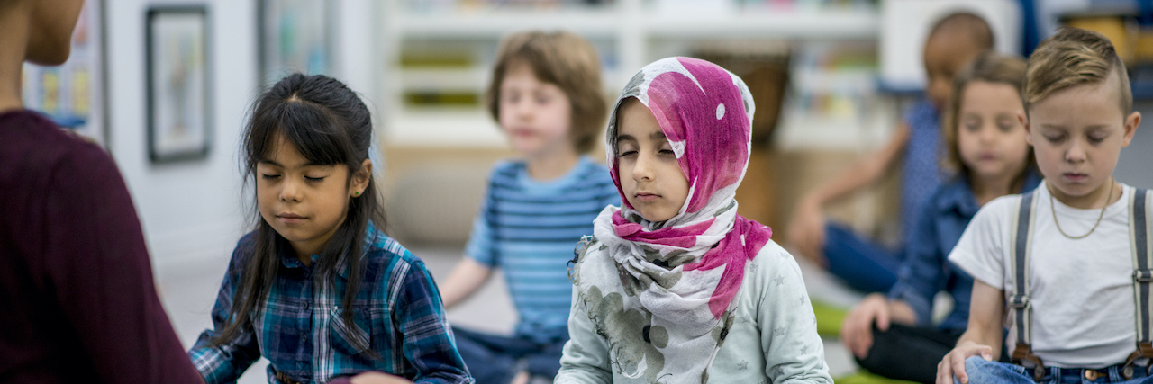The Mental Health and Academic Benefits of Mindfulness for Children A multi-ethnic group of young school children are sitting on pillows with their eyes closed and hands on their laps