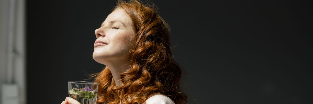 Coming to Terms With an Abusive Relationship and My Bipolar Disorder A woman with red, curly hair stands in the sunlight holding a glass of water with a floral garnish.