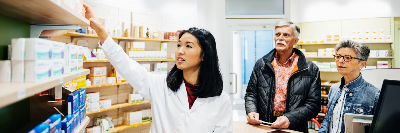 Why I'm Thankful for Pharmacy Staff as Someone With Chronic Illness A pharmacist helping a mature couple fill prescriptions at local pharmacy..