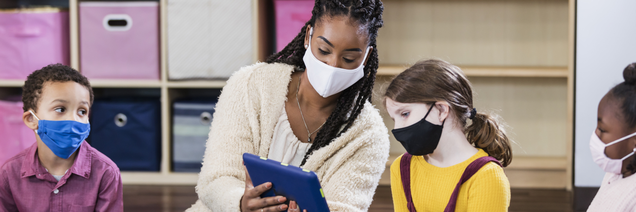 School Mask Mandates Are a Disability Rights Issue A multi-ethnic group of kindergarten or first grade students and their teacher in the classroom, sitting together in a circle on the floor. The children are 5 to 7 years old. They are all wearing masks, back to school during the COVID-19 pandemic, trying to prevent the spread of coronavirus. The teacher, an African-American woman, is holding a digital tablet up, showing it to the girl sitting next to her.