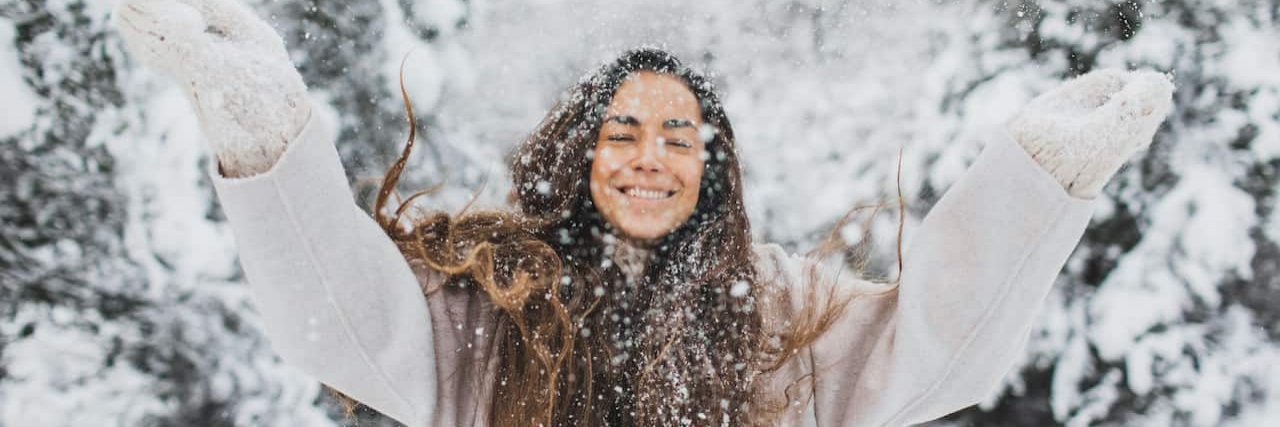 Woman standing in snowy landscape with long brown hair smiling and throwing snow in the air