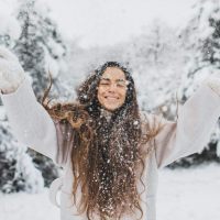 Woman standing in snowy landscape with long brown hair smiling and throwing snow in the air