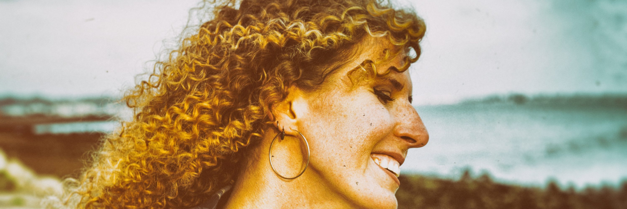 Smiling woman with curly hair and headphones around her neck, walking outdoors.