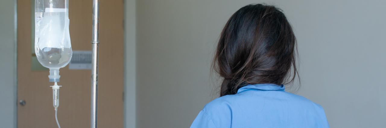 What It's Like Being Young With Crohn's Disease the back view of a younger woman in a hospital walking with an IV machine