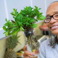 A Black woman wearing a gray sweater and holding a plant in a jar smiles for the camera.