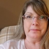 A close-up photo of a white woman with long blonde hair wearing glasses and a white shirt.
