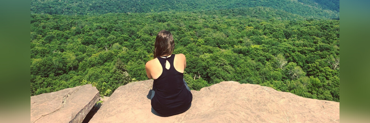 How Anxiety and Depression Make Me Believe I'm Too Broken to Be Loved Photo of author; back of a young woman sitting on a rock overlooking mountains and trees