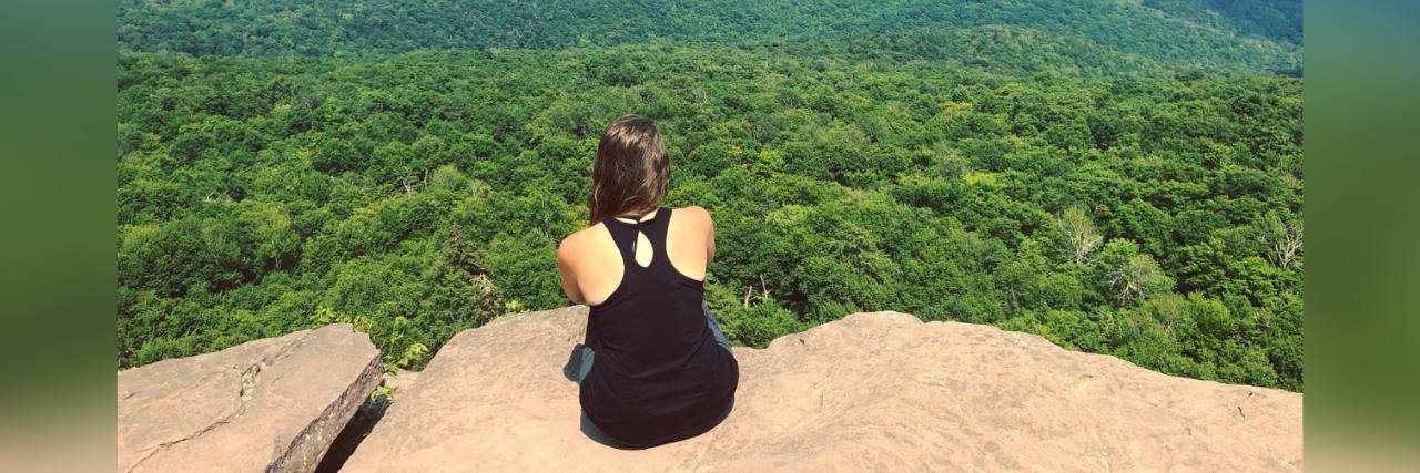 Photo of author; back of a young woman sitting on a rock overlooking mountains and trees