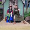 Two nerds sit together looking cute at a renaissance fair. The partner on the left has blue hair and wears a red shirt, blue skirt, and green overlap. On the right hand side the other partner wears a santa hat, and a green elven costume with brown boots. He has a wooden staff in his hands. They're both smiling. they're really cute.