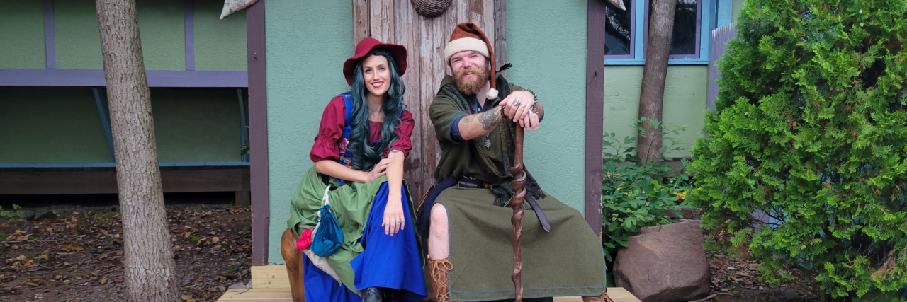 What it's Like to Start a New Relationship With Cancer Two nerds sit together looking cute at a renaissance fair. The partner on the left has blue hair and wears a red shirt, blue skirt, and green overlap. On the right hand side the other partner wears a santa hat, and a green elven costume with brown boots. He has a wooden staff in his hands. They're both smiling. they're really cute.
