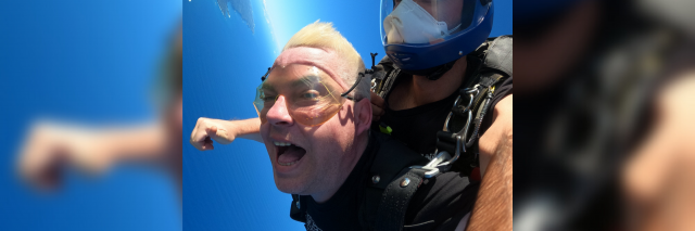 Two men are attached to one another jumping out of an airplane. One man is definitely scared out of his life, the other looks up at the camera. Blue ocean and green forrest is below them