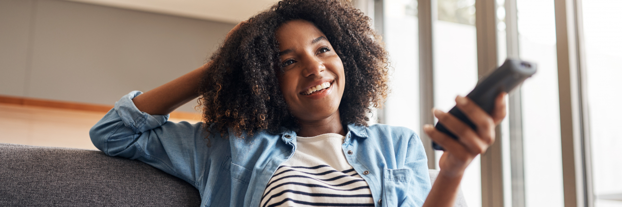 A Black woman with brown eyes and natural hair wearing a white-and-black striped shirt and a denim jacket holds up a remote control while smiling.