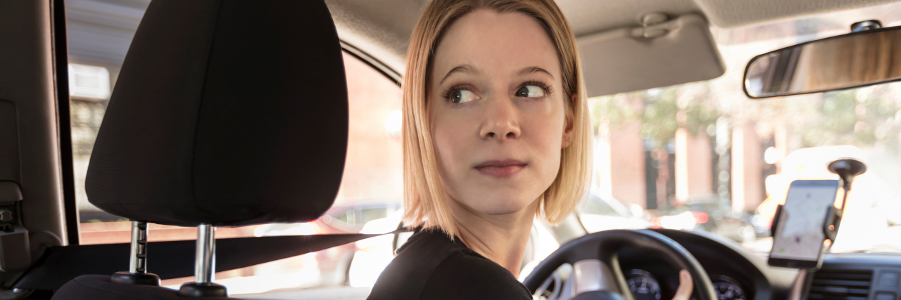 Being Publicly Humiliated During a Depressive Episode A woman with short blonde hair and brown eyes wearing a black T-shirt looks behind her while sitting behind the wheel of a car. Her hand is on the steering wheel.