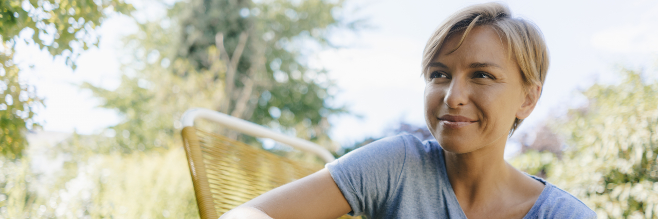 Dreaming New Dreams During Loss of Plans From Schizoaffective Disorder A woman with short blonde hair and brown eyes sits outside on a wicker chair while holding a book in her hands.