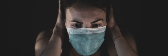 Woman in a blue surgical mask with her hands over her ears, looking distressed