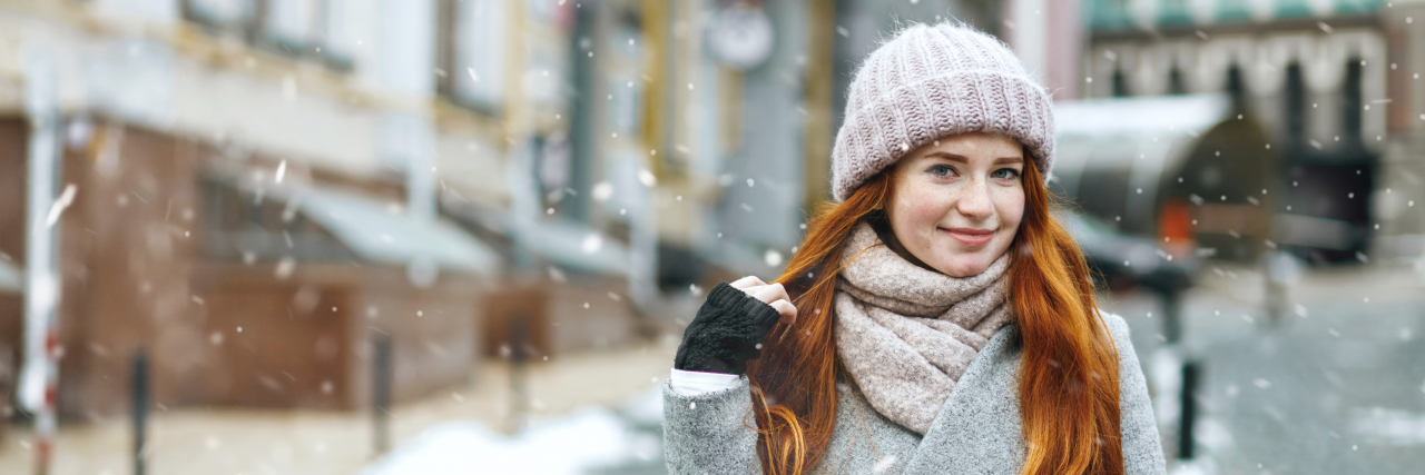 A young red-haired woman wearing a gray winter coat poses confidently as snow falls around her.